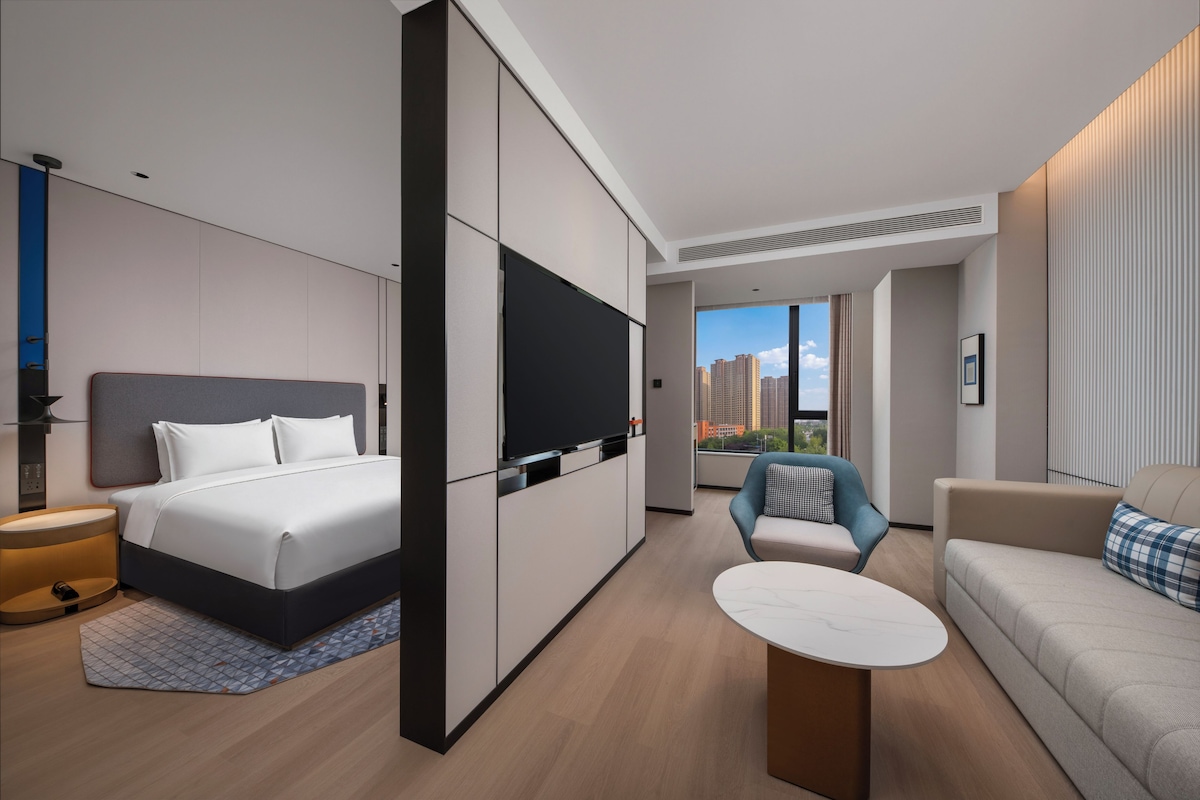 A modern hotel room with a bed, a TV, a seating area with a table, and a view of the cityscape.