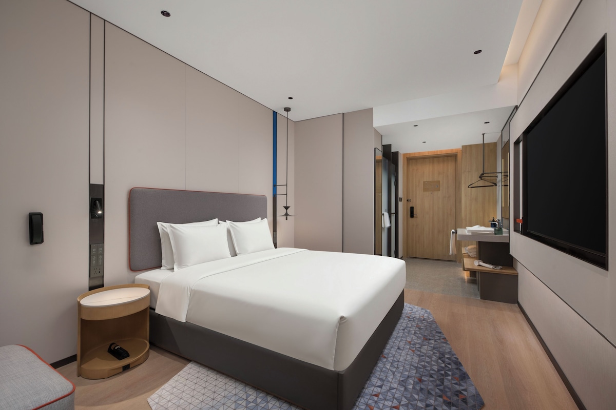 A modern hotel room with a large bed, a bedside table, a small round table, a mirror, a TV, and a bathroom with a sink and shower.