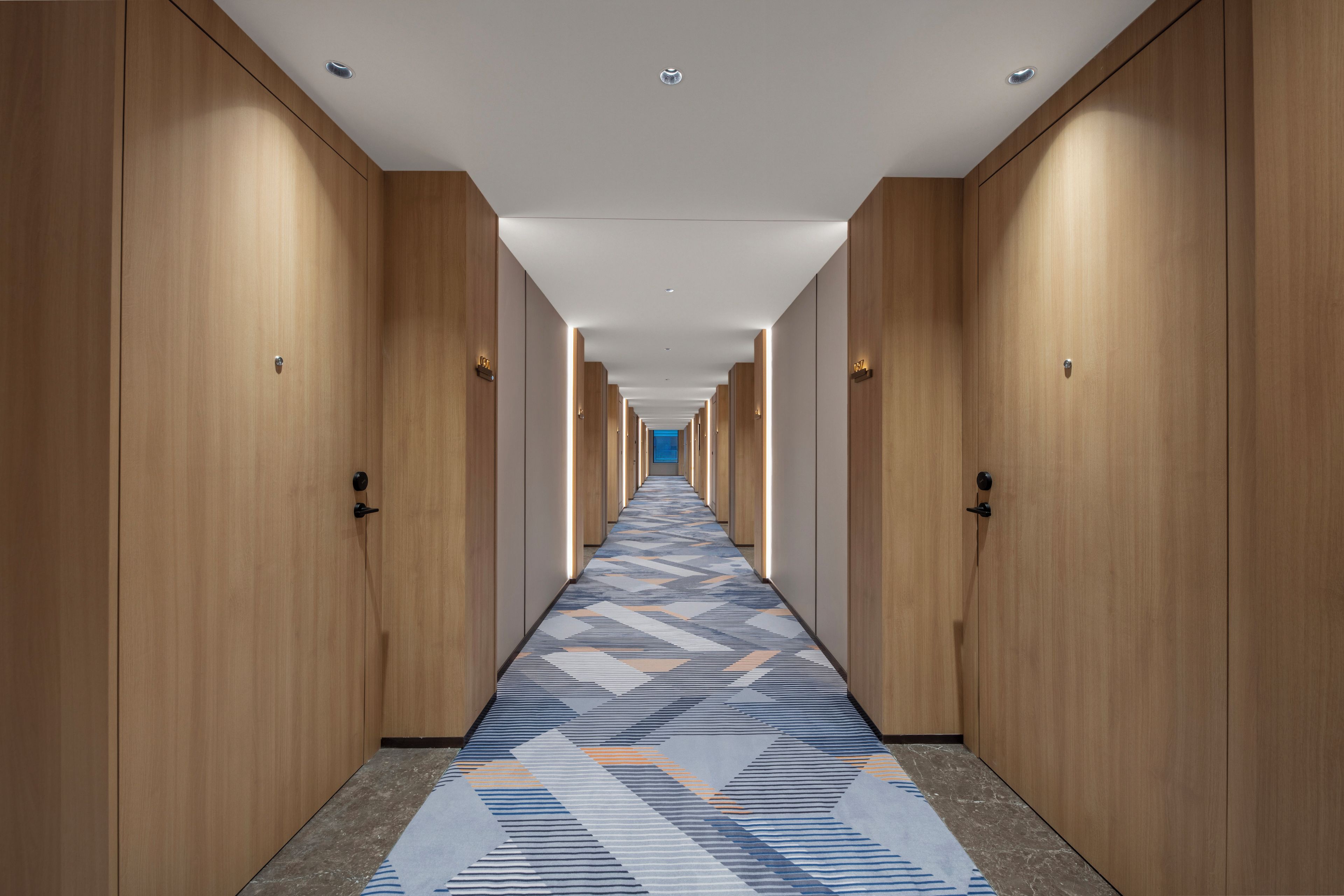 A hotel corridor with wooden doors, a patterned carpet, and a straight ceiling with recessed lighting.