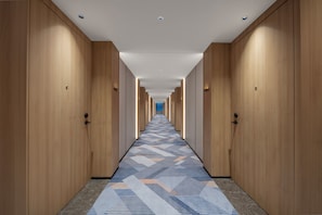 A hotel corridor with wooden doors, a patterned carpet, and a straight ceiling with recessed lighting.