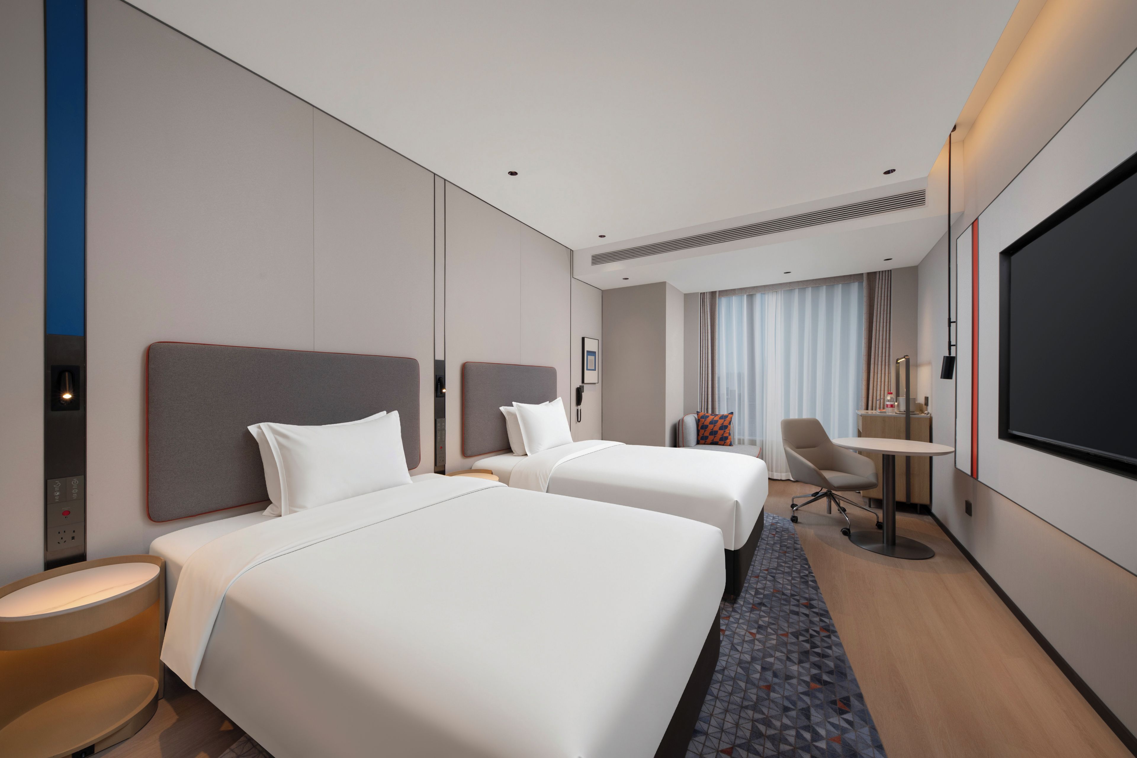 A hotel room with two beds, a desk, a chair, a TV, and a large window with curtains.