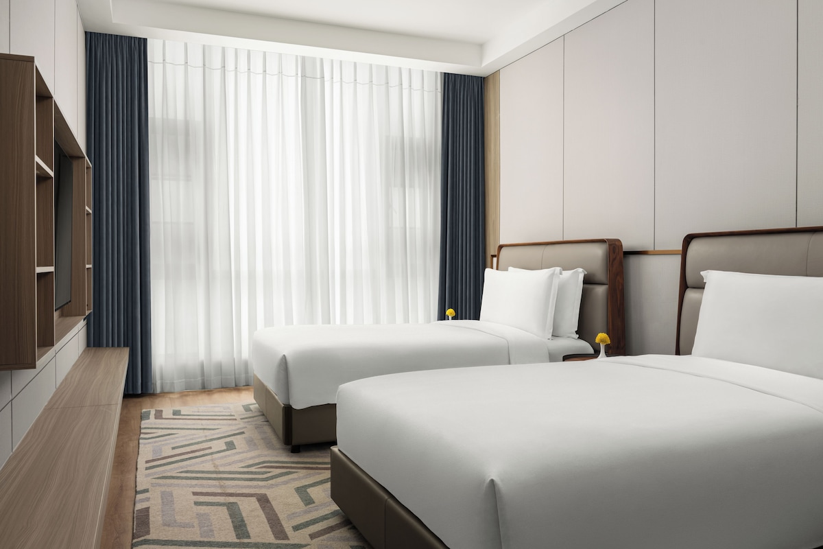 A hotel room with two beds, a TV, and a large window with curtains.