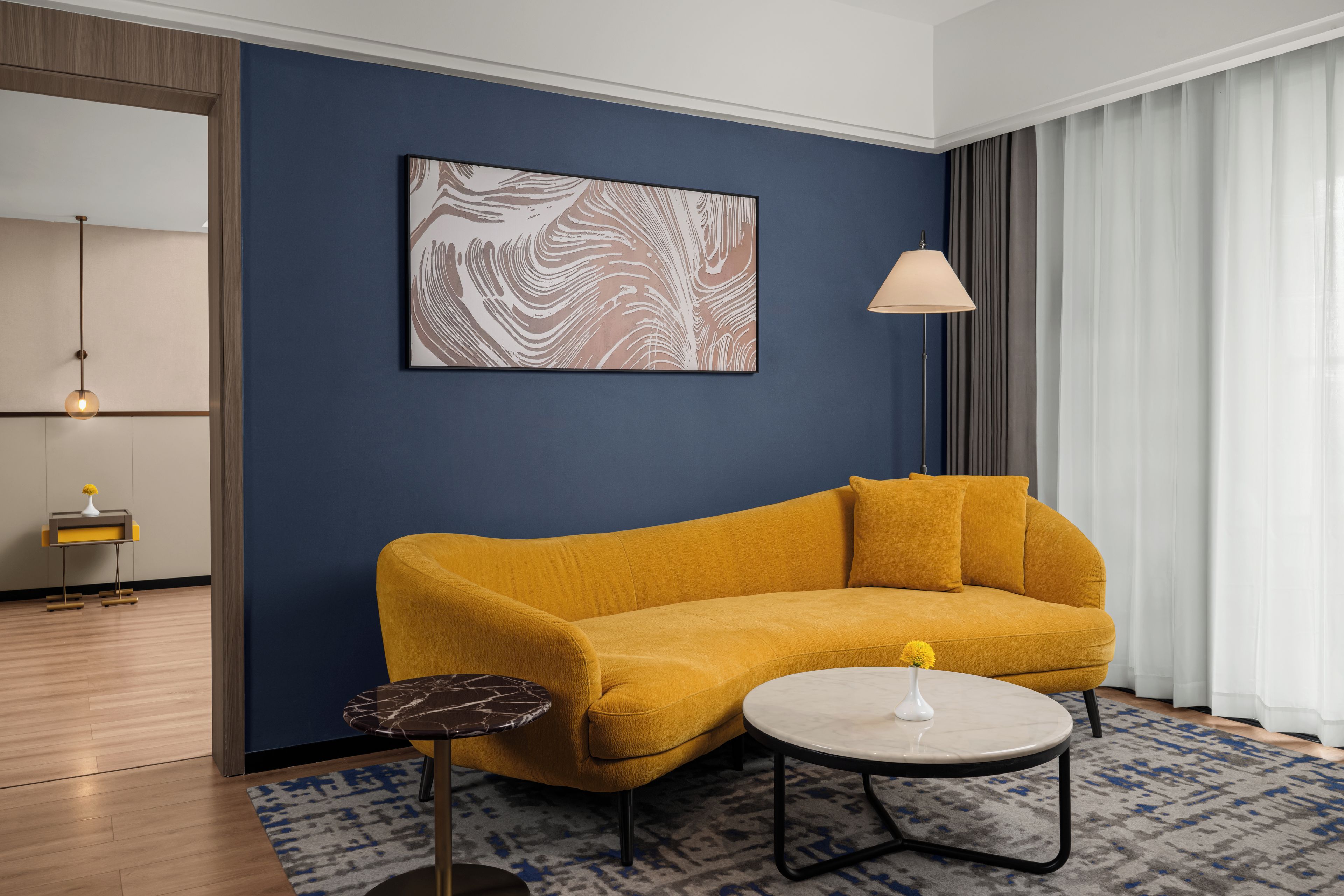 Una sala de estar moderna con una pared azul, un sofá color mostaza, una mesa de centro redonda, una lámpara de pie y una mesita auxiliar minimalista.