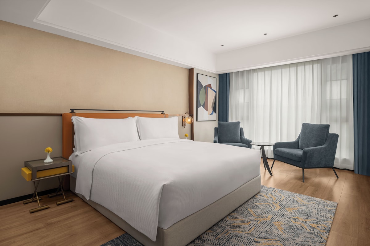 A hotel room with a large bed, a desk, a chair, and a large window with curtains.
