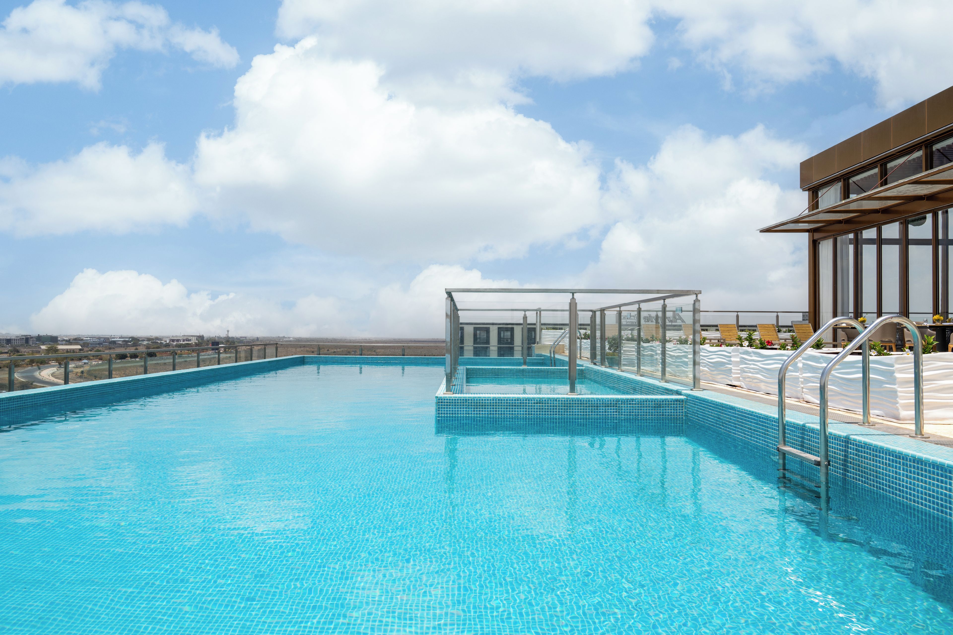 Una piscina azul cristalina con cerramiento de vidrio y escalones de acceso, rodeada por un edificio moderno con amplios ventanales.