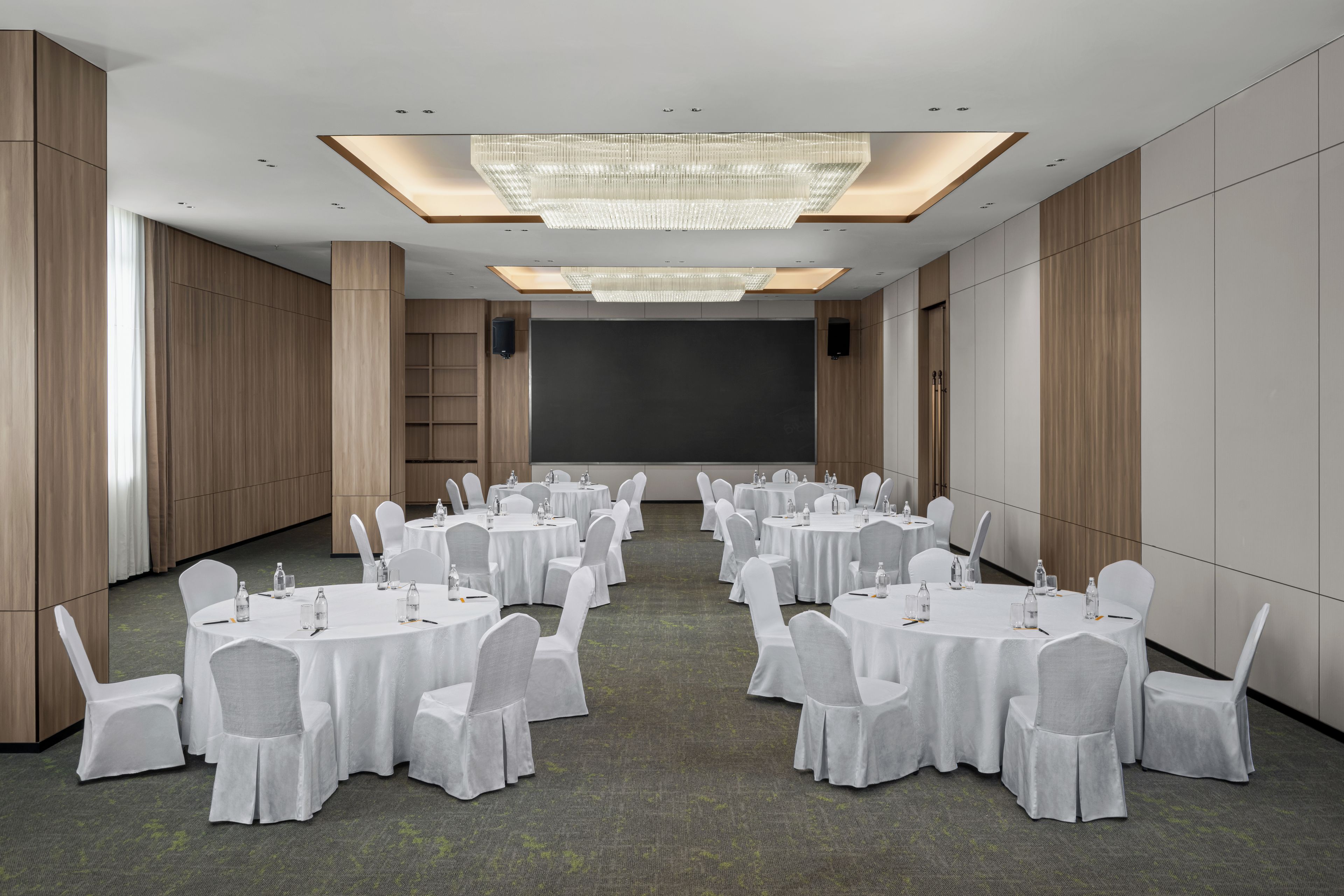 Un salón de conferencias con mesas redondas, sillas blancas, una pantalla grande y una araña.