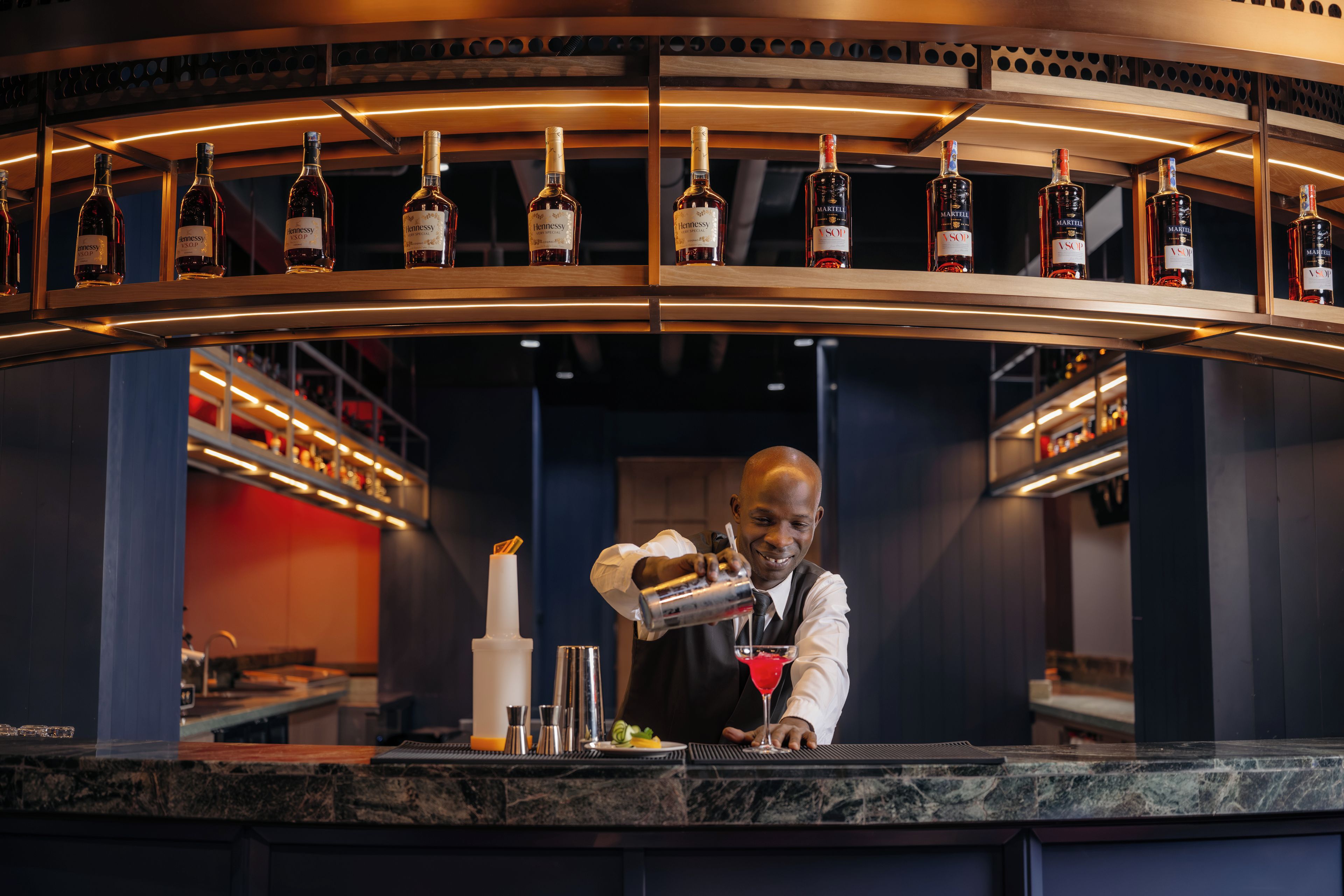 Zona de bar con un bartender preparando una bebida, estanterías llenas de botellas de licor y una encimera de mármol.