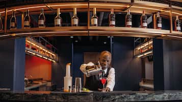 Zona de bar con un bartender preparando una bebida, estanterías llenas de botellas de licor y una encimera de mármol.