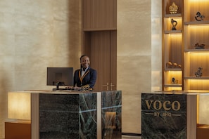 A reception desk with a man standing behind it, a wooden panel wall, and a shelf with various items.