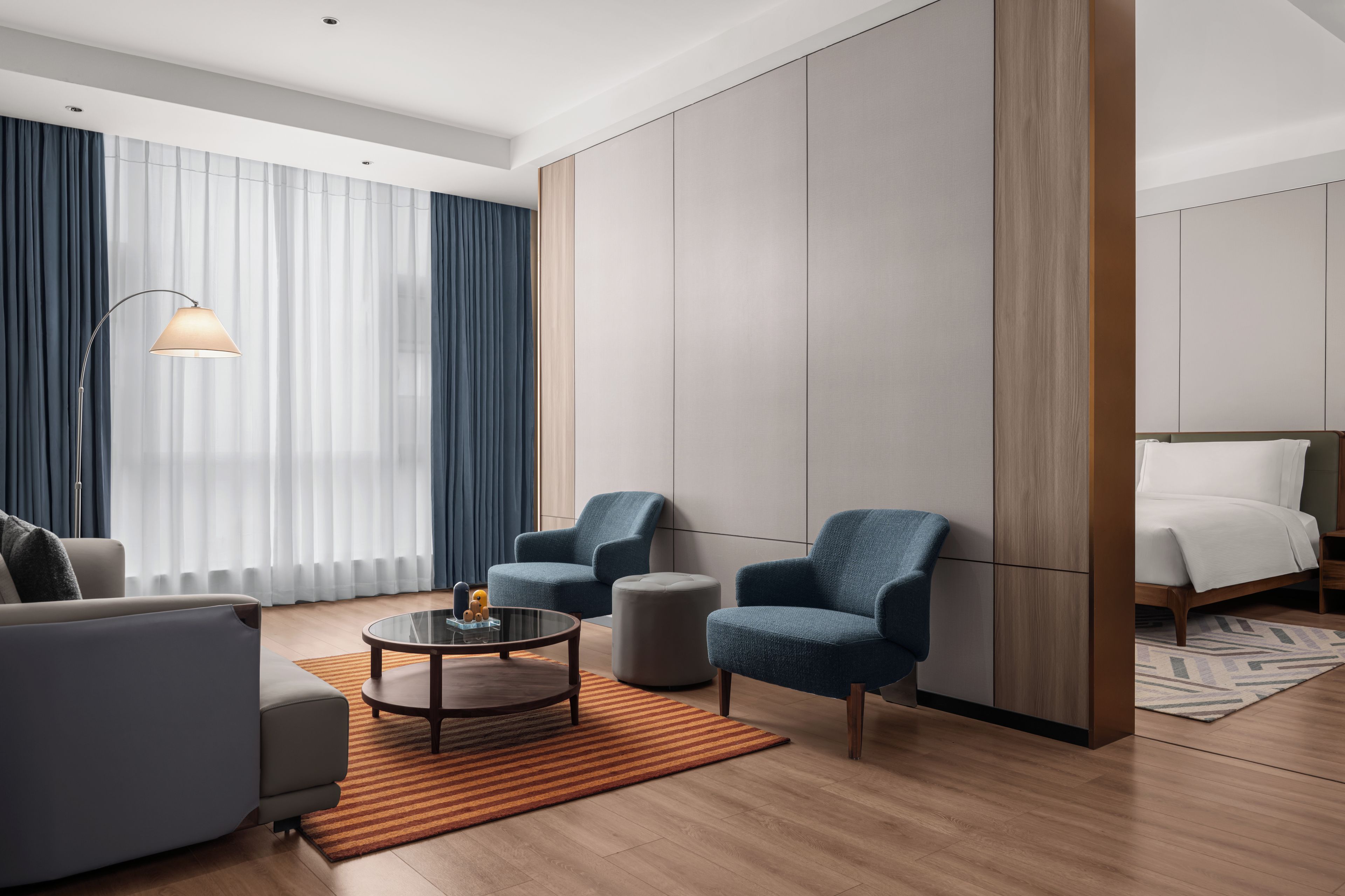 Una habitación de hotel moderna con sofá, sillones, una mesa de centro, cama y un gran ventanal con cortinas.