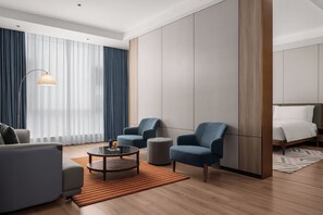A modern hotel room with a sofa, armchairs, a coffee table, a bed, and a large window with curtains.