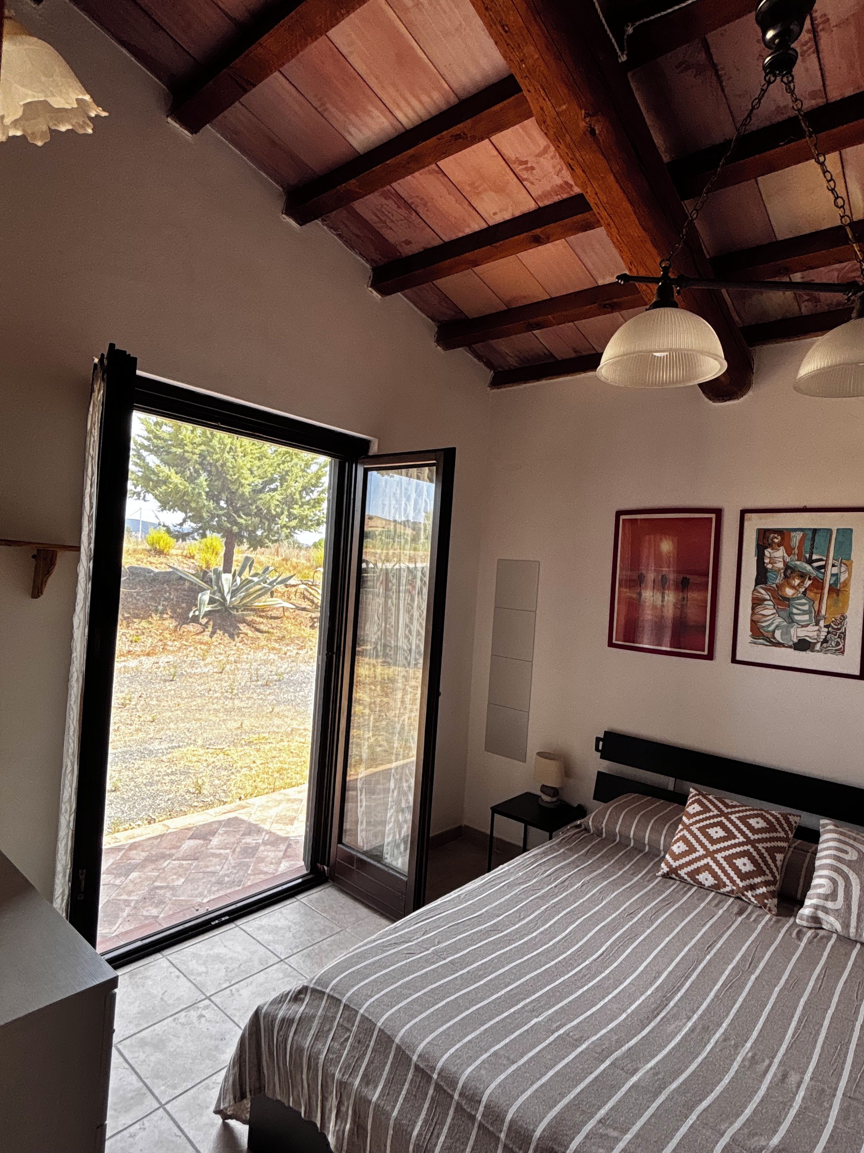 Un dormitorio con cama, mesita de noche, lámpara, ventana con vista a árboles y ventilador de techo.
