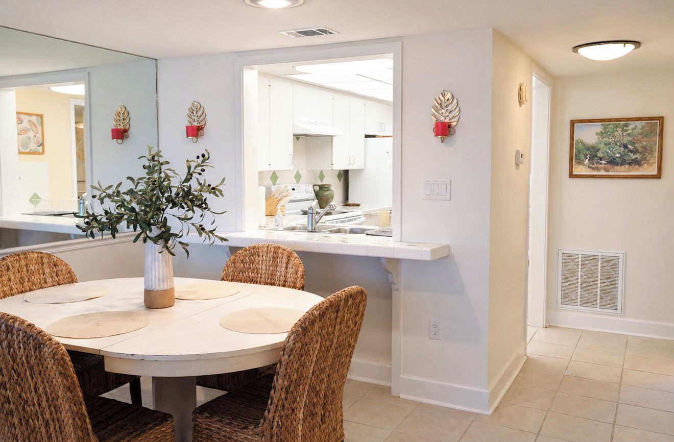 A dining area with a round table, wicker chairs, a vase with greenery, and a kitchen visible through an open doorway.