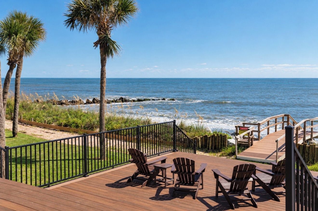 Een houten terras met stoelen, met uitzicht op een strand met palmbomen en een rotsachtige kustlijn.