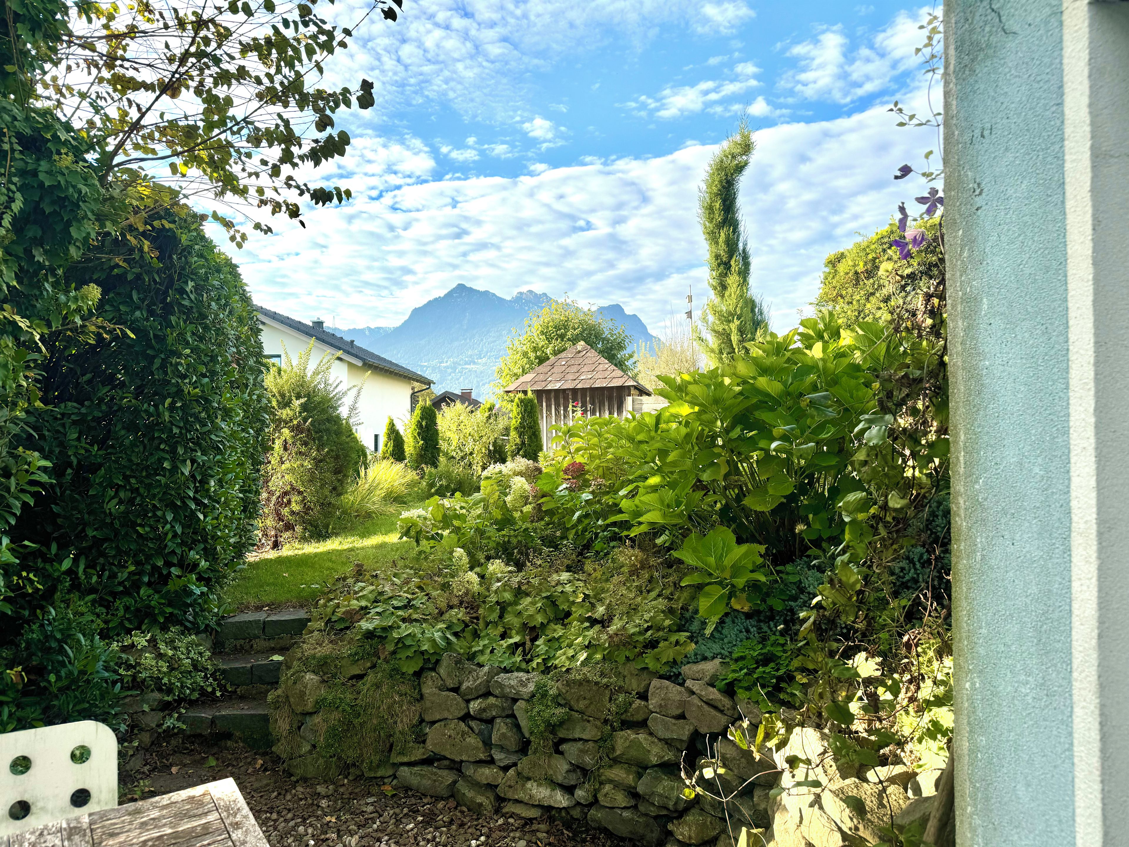 Un jardin avec un mur de pierre, un banc en bois et une vue sur des montagnes au loin.