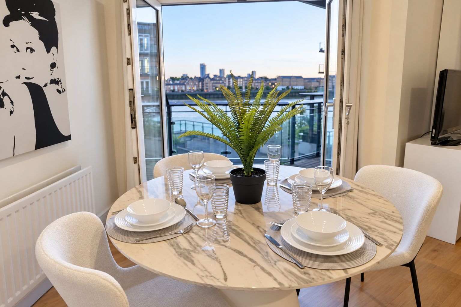 A dining area with a table set for four, a potted fern, and a city view through a glass door.