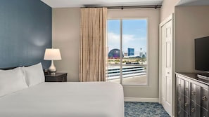 Une chambre d’hôtel avec un grand lit, une table de chevet avec une lampe, un téléviseur posé sur une commode et une vue sur le paysage urbain, visible à travers une fenêtre avec des rideaux.