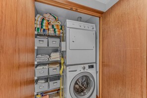 A laundry room with a washing machine and dryer, shelves with folded towels, and storage bins. - Cozy Condo at Waterville Estates (Thornton)