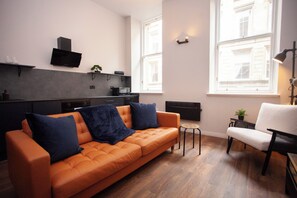 Ein modernes Wohnzimmer mit einem orangefarbenen Sofa, einem weißen Sessel und einem kleinen runden Tisch. Zwei Fenster sorgen für natürliches Licht, und es gibt eine Küchenzeile mit schwarzer Arbeitsplatte und Schränken.
