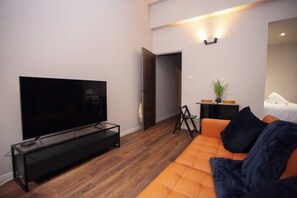 En moderne stue med et fladskærms-tv på et glasbord, en orange sofa med mørke puder og et lille sofabord med en vase.