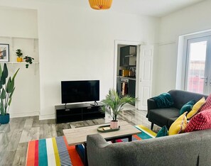 Ein Wohnzimmer mit einem Flachbildfernseher, einem grauen Sofa mit bunten Kissen, einem Holztisch und einem Streifen Teppich.