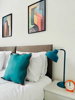 Ein ordentlich bezogenes Bett mit weißer Bettwäsche und einem türkisfarbenen Kopfkissen, eine blaue Schreibtischlampe und ein klassischer orangefarbener Wecker auf einem Nachttisch.