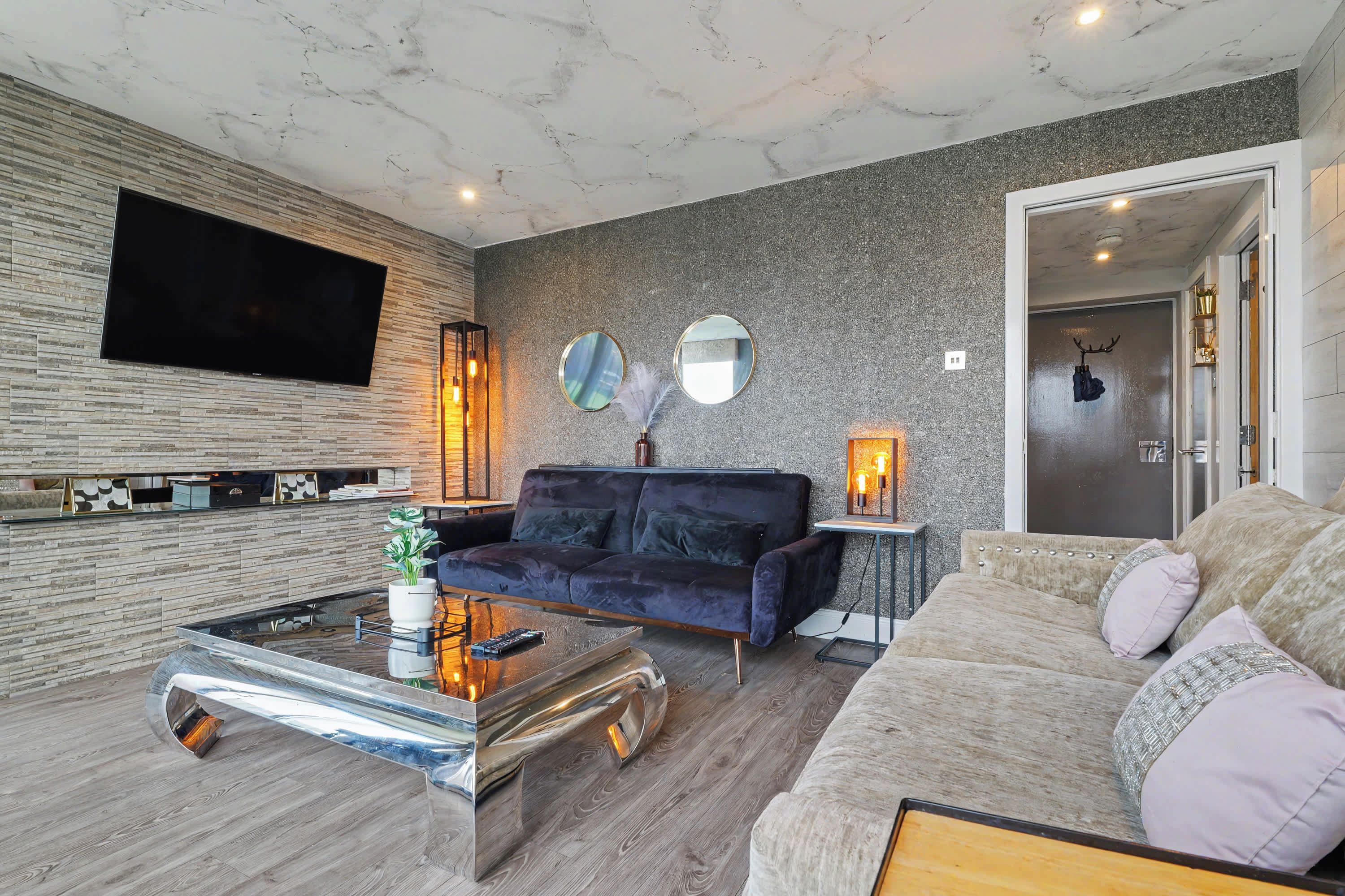 Una sala de estar moderna con un televisor de pantalla plana, un sofá azul oscuro, una mesa de centro de vidrio y una pared gris con textura.