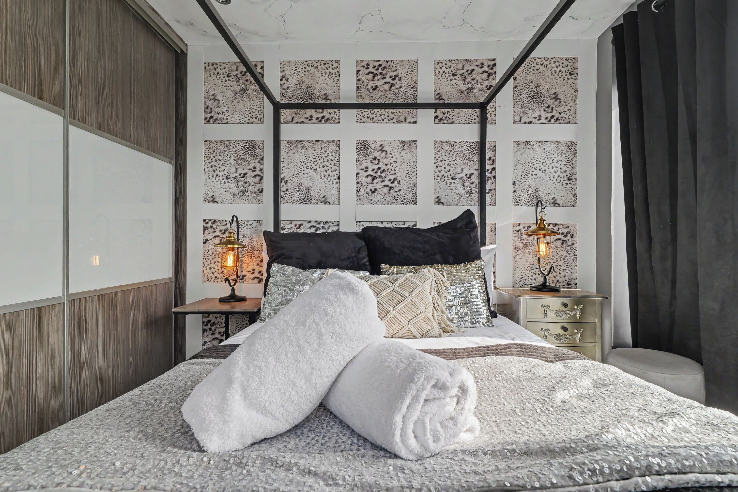 Een moderne slaapkamer met een hoofdbord met een geometrisch patroon, een groot bed met witte handdoeken, twee bedlampjes en een nachtkastje met een decoratief object.