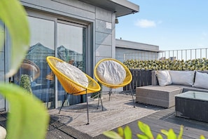A rooftop terrace with two yellow wicker chairs, a grey sofa, and a glass door leading to a building.