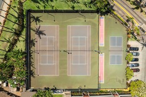 Ein Tennisplatzkomplex mit mehreren Plätzen, umgeben von Grünflächen und neben einer Straße mit geparkten Autos.