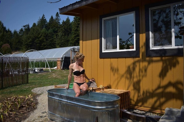 A person in a bathtub outside a yellow building with a window, surrounded by a garden and a greenhouse.