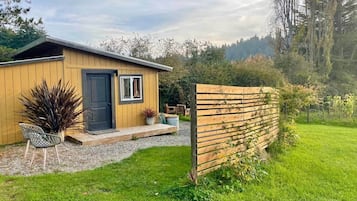 A wooden cabin with a door and windows, a gravel path leading to the door, and a grassy yard with a wooden fence.