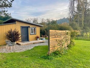 Une cabane en bois avec une porte et des fenêtres, un chemin de gravier menant à la porte et une cour herbeuse bordée d’une clôture en bois.
