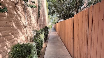 A narrow concrete pathway flanked by a brick wall on one side and a wooden fence on the other, leading to a building entrance.