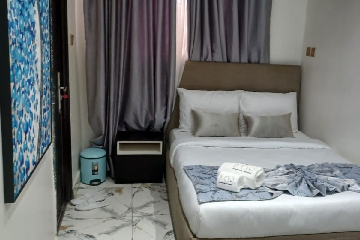 Une petite chambre à coucher, bien rangée, avec un lit, une table de chevet, une poubelle et une porte munie d’un rideau.