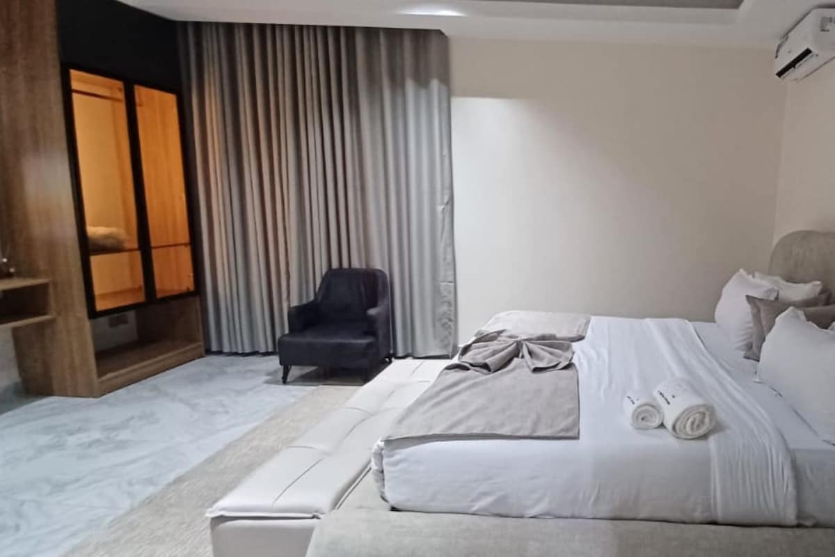 Une chambre d’hôtel moderne avec un grand lit, une chaise et une vue sur une pièce dont les fenêtres sont fermées par des rideaux.