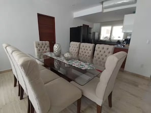 A dining area with a glass-top table, four upholstered chairs, and a kitchen in the background. - Acapulco Diamante / jacuzzi / club de playa / 2 px (Acapulco de Juárez)