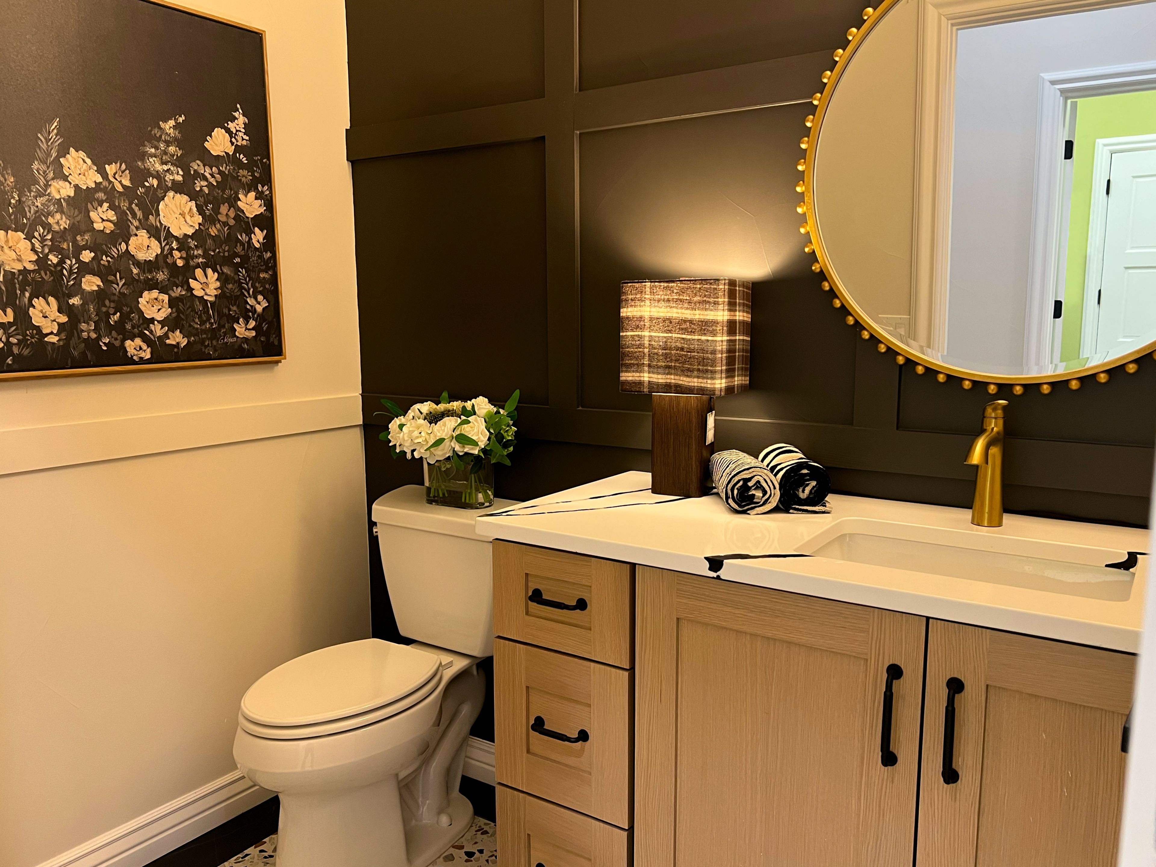 A bathroom with a toilet, a sink with a mirror, a lamp, and a framed floral artwork on the wall.