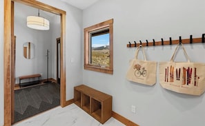 Ein Flur mit einer Holzbank, einem Spiegel, einer Pendelleuchte, einem Fenster mit Landschaftsblick und zwei Stofftaschen auf einem Gestell.