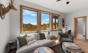 Una sala de estar moderna con sofá, sillón y reposapiés, una cabeza de venado de madera montada en la pared, y ventanas grandes que ofrecen vistas al paisaje.