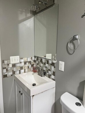 A bathroom with a white sink, a mirror above it, and a toilet. There are two electrical outlets on the wall and a soap dispenser on the sink.