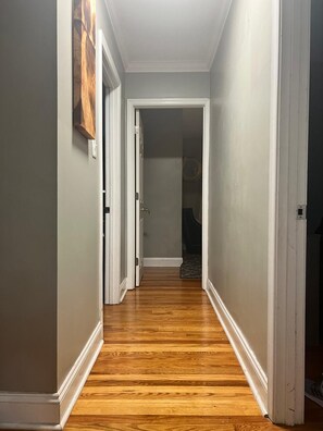 A hallway with wooden flooring and white trim, leading to a room with a patterned rug.
