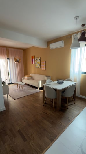 A modern living room with a sofa, dining table, and chairs. There is a ceiling fan, an air conditioning unit, and a window with curtains. - Apartments Sava (Tivat)