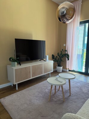 Un saló amb una televisió plana, un armari blanc, dues taules rodones blanques i plantes en testos.
