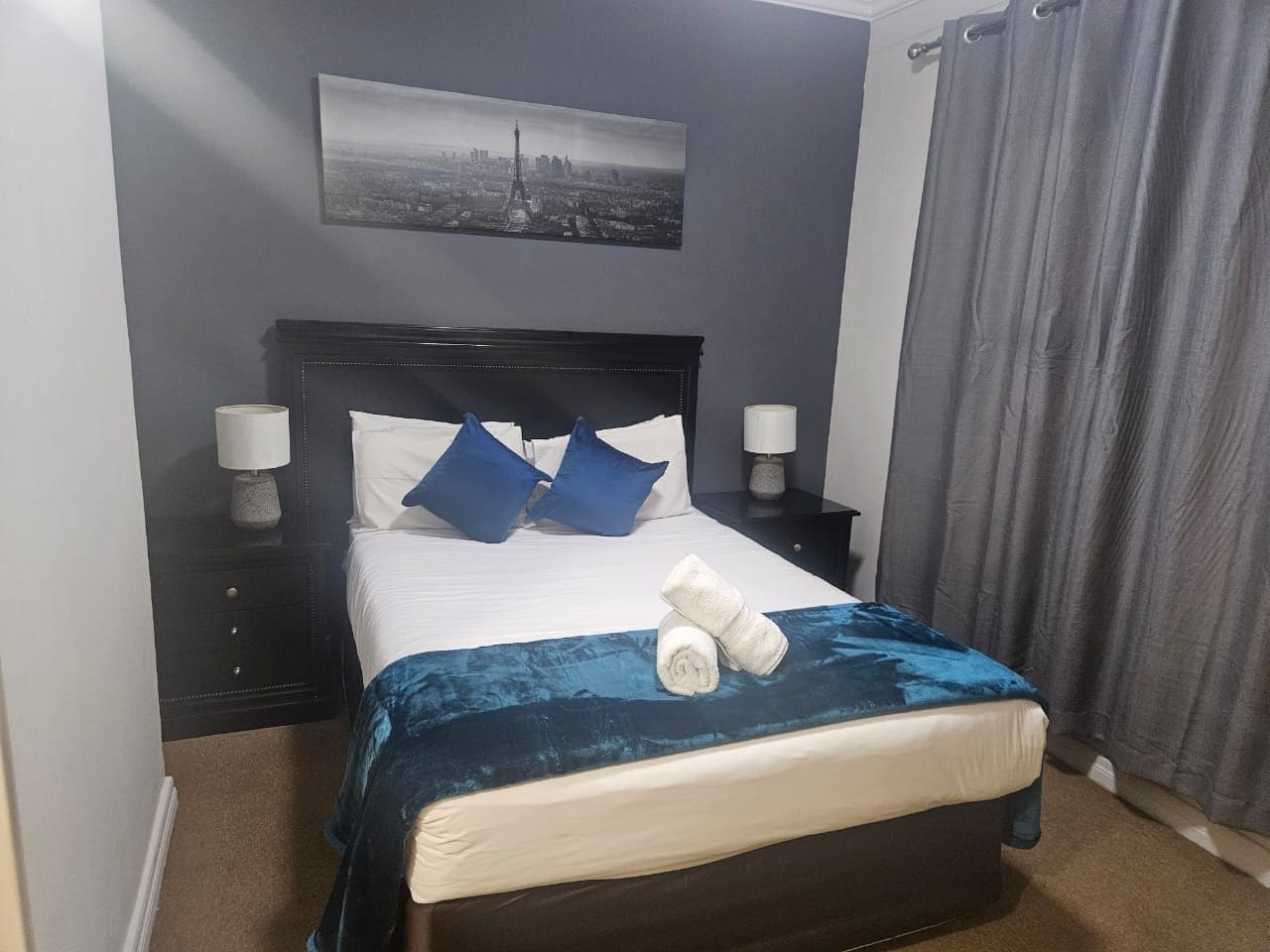 Una cama bien hecha con una manta azul, dos almohadas y toallas dobladas. Se ve una mesita de noche con una lámpara y un cuadro en la pared.
