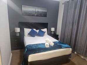 A neatly made bed with a blue blanket, two pillows, and rolled towels. There is a nightstand with a lamp and a wall-mounted picture. - Sandton Westpoint Luxury Apartments 158 (Sandton)
