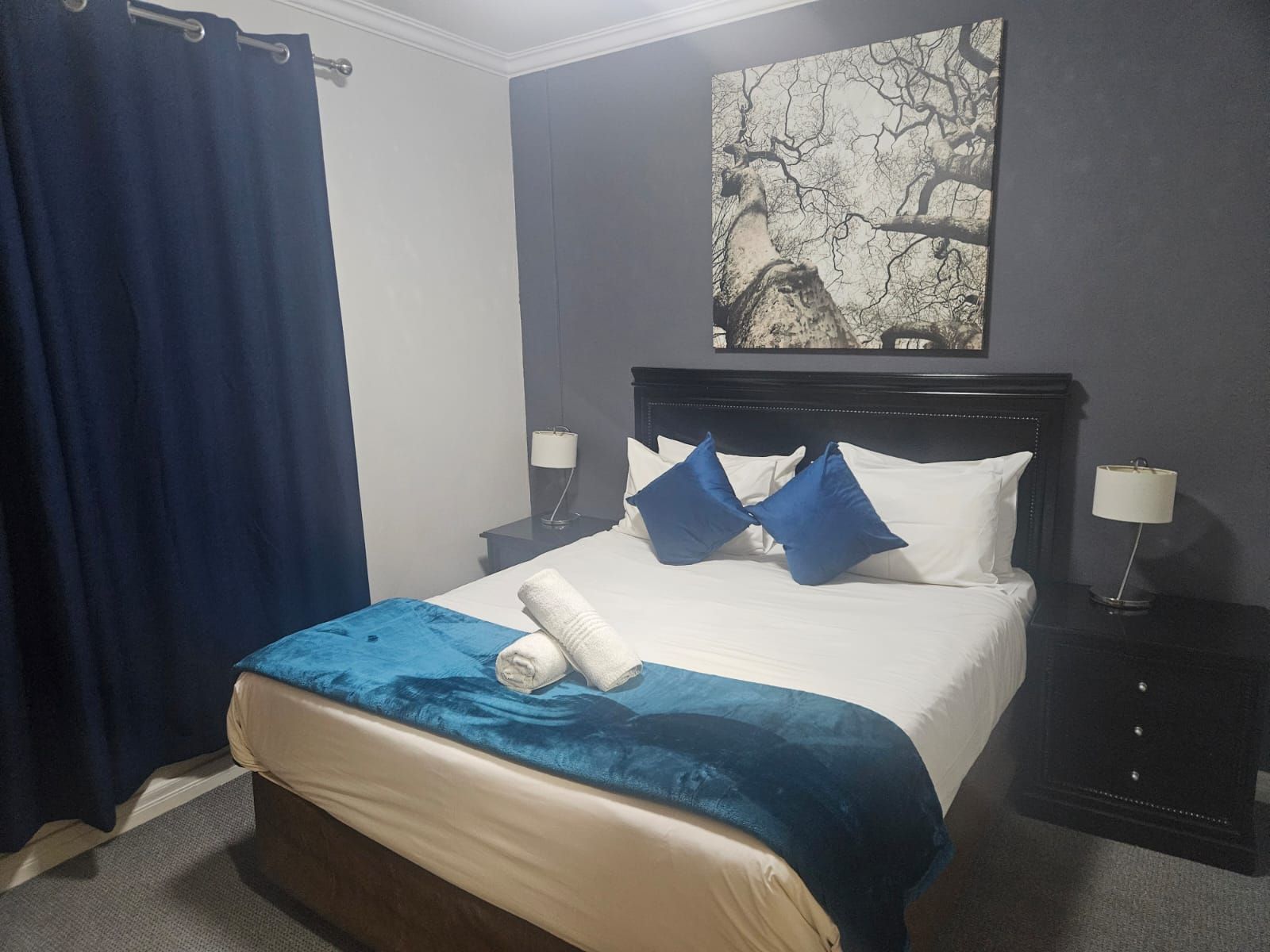 Una habitación de hotel con cama, mesita de noche, lámpara, cortina azul y una obra de arte en la pared.