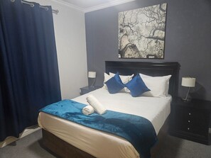 Quarto de hotel com cama, mesa de cabeceira, luminária, cortina azul e uma obra de arte na parede.