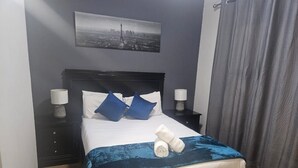 Una cama bien hecha con una manta azul, dos almohadas y toallas dobladas. Se ve una mesita de noche con una lámpara y un cuadro en la pared.