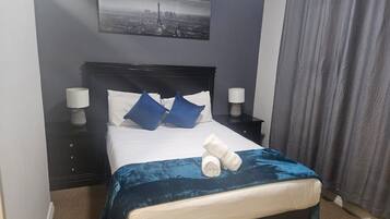 A neatly made bed with a blue blanket, two pillows, and rolled towels. There is a nightstand with a lamp and a wall-mounted picture.